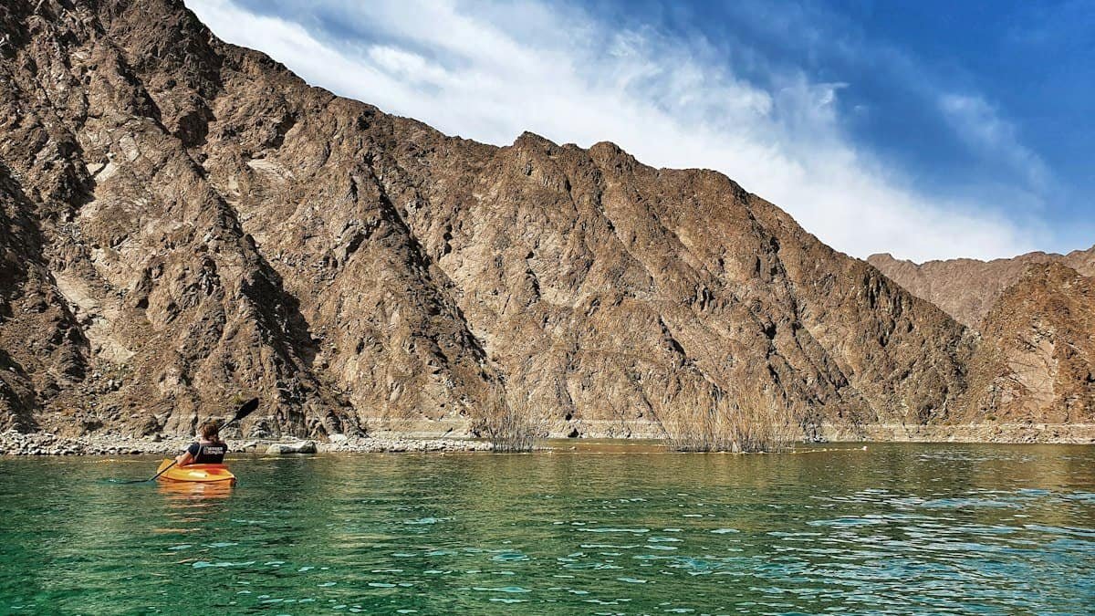A lone kayaker in an orange kayak on green water below steep rocky mountains