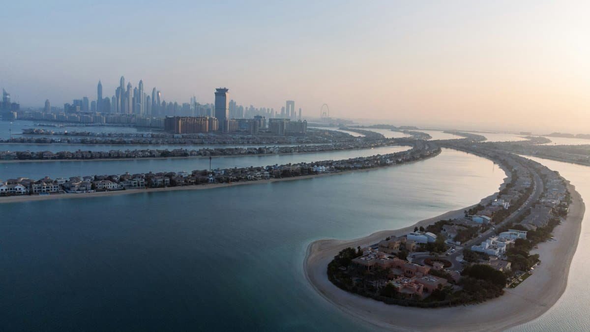 Aerial view of Palm Jumeirah fronds lined with villas at dusk, with the Dubai Marina skyline beyond